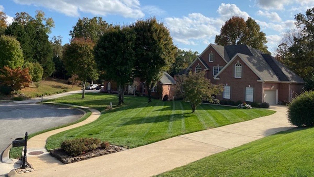 Outdoor Inc maintained estate lawn with striped turf, mature shade trees, and a brick home