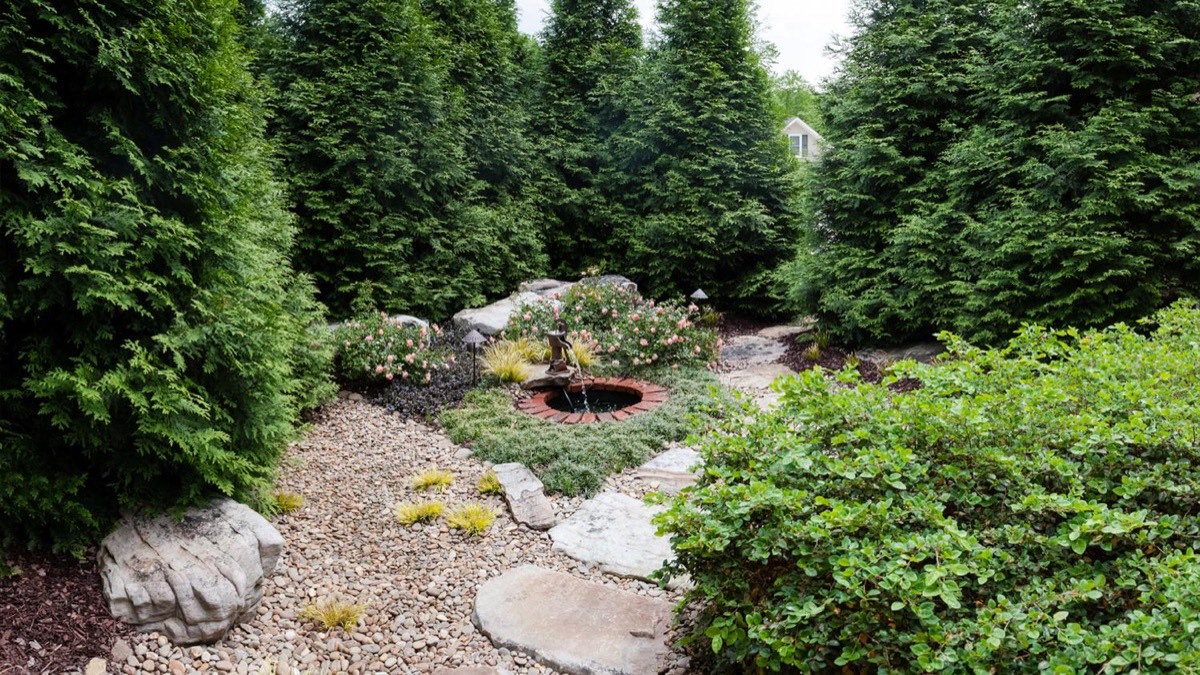 Outdoor Inc garden path with boulders, gravel, roses, and a fountain framed by evergreens